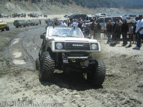 1987 Suzuki Samurai - A modified Suzuki Samurai navigates a muddy terrain near a body of water, with spectators gathered.