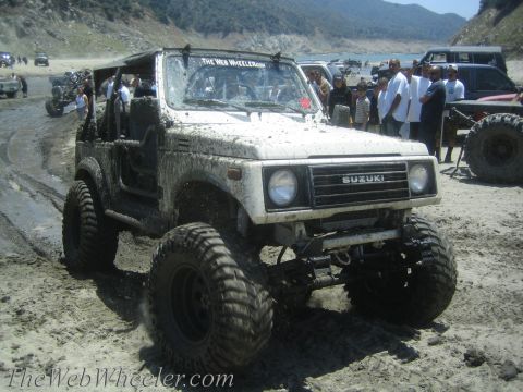 1987 Suzuki Samurai - A white 1987 Suzuki Samurai, covered in mud, navigates a sandy and wet terrain near a body of water with other vehicles and people present.