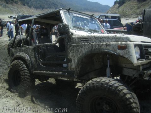 1987 Suzuki Samurai - A heavily mud-splattered 1987 Suzuki Samurai navigates a sandy, off-road terrain with other vehicles in the background.
