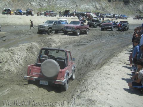 1987 Suzuki Samurai - A group of off-road vehicles, including a red Suzuki Samurai, navigate a muddy track with onlookers.