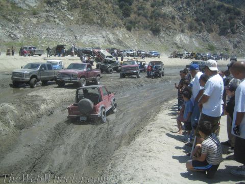 1987 Suzuki Samurai - A group of off-road vehicles are seen navigating a muddy terrain with onlookers observing the action.