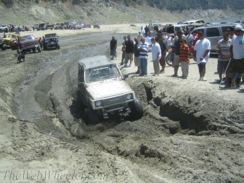 1987 Suzuki Samurai - A group of people watch as a Suzuki Samurai tackles a challenging muddy terrain on a beach.