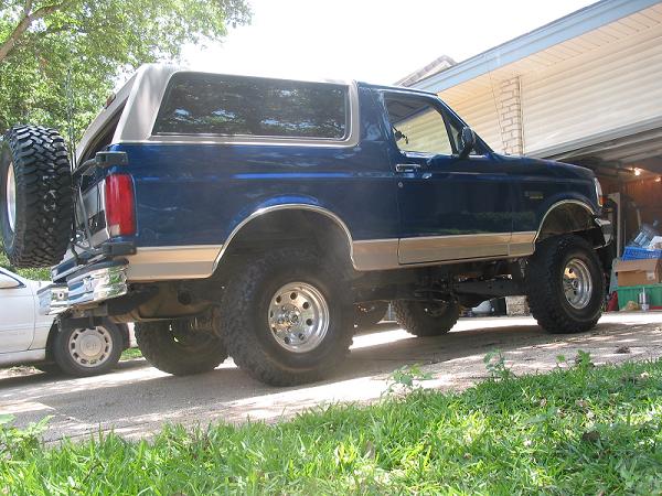 Lifted blue and tan Ford Bronco with grey top parked by a garage, rear three-quarter