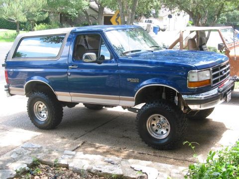1996 Ford Bronco - A lifted blue Ford Bronco with tan side trim and aftermarket wheels is parked on a concrete driveway.
