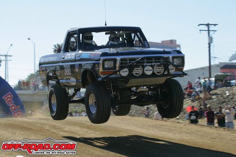 1990 Ford Bronco - black vintage Ford Bronco race truck numbered 300 catches air over a dirt course as spectators line the background.