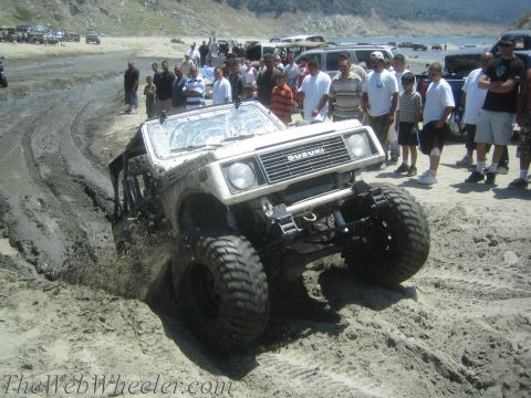 1987 Suzuki Samurai - A white 1987 Suzuki Samurai navigates a muddy track with a crowd gathered to watch the off-road action.