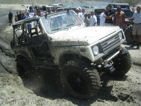 1987 Suzuki Samurai - Mud-covered 1987 Suzuki Samurai navigates a sandy, wet terrain with spectators watching in the background.