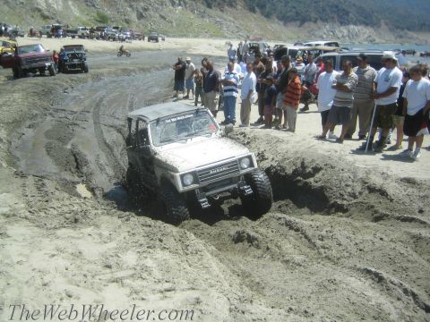 1987 Suzuki Samurai - A white 1987 Suzuki Samurai tackles a muddy obstacle on a beach, with a crowd of onlookers watching the off-road action.