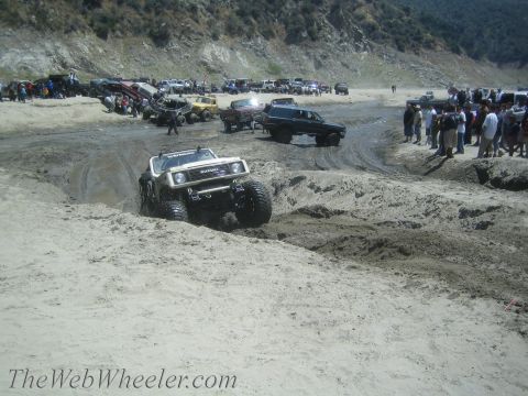 1987 Suzuki Samurai - A modified 1987 Suzuki Samurai tackles a challenging muddy terrain, with other vehicles and spectators in the background.