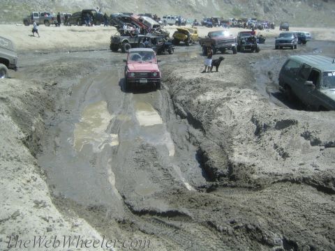 1987 Suzuki Samurai - A red 1987 Suzuki Samurai navigates a challenging muddy obstacle course with other vehicles in the background.