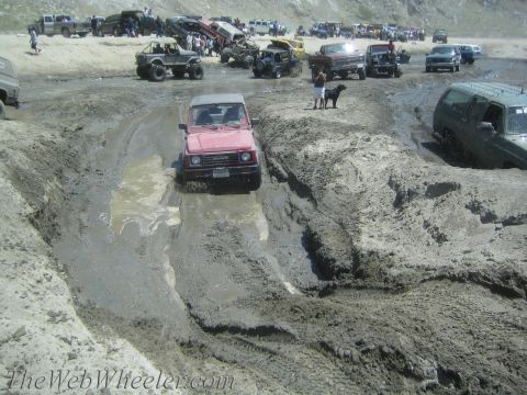 1987 Suzuki Samurai - A red 1987 Suzuki Samurai tackles a challenging mud pit while other off-road enthusiasts watch and participate in the background.