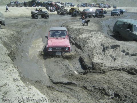 1987 Suzuki Samurai - Red Suzuki Samurai navigating a muddy track with spectators and other off-road vehicles nearby.