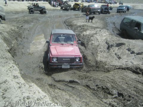1987 Suzuki Samurai - A red Suzuki Samurai navigates a muddy off-road course with other vehicles and spectators in the background.