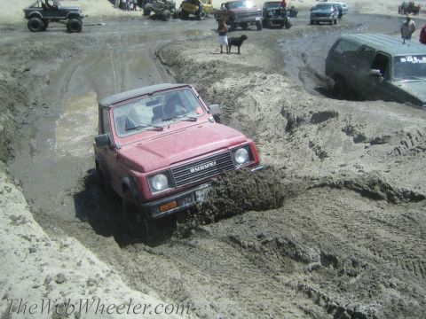 1987 Suzuki Samurai - A red 1987 Suzuki Samurai navigates a muddy terrain, with other off-road vehicles visible in the background.