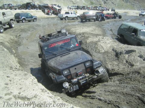 1987 Suzuki Samurai - Multiple off-road vehicles navigate a muddy terrain, with one Jeep Wrangler prominently stuck in a deep mud pit.