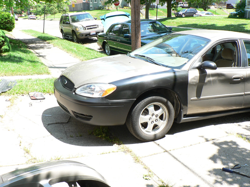 Gray Ford Taurus sedan in driveway with flat black painted front bumper and fender, parts on ground nearby