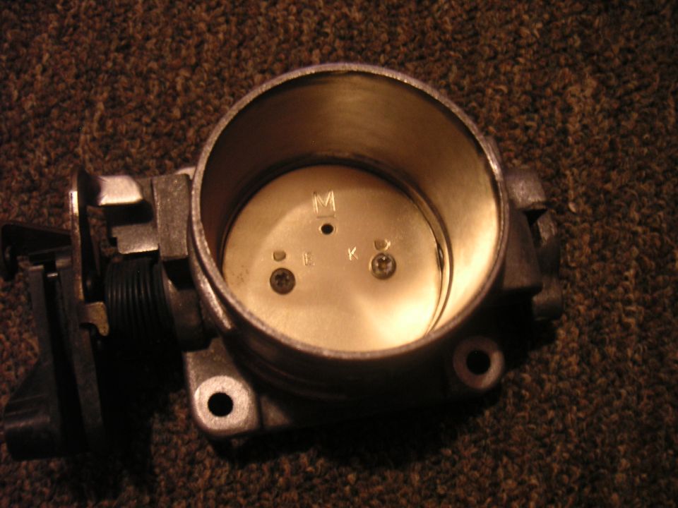 Throttle body with markings M, L, K, and D. It is mounted on a dark, textured surface