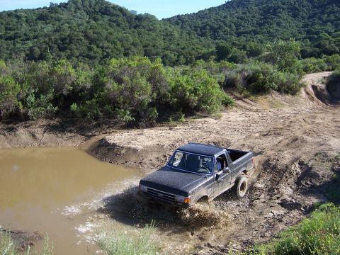1991 Ford Bronco - A dark pickup truck navigates a muddy obstacle on an off-road trail.