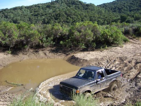 1991 Ford Bronco - A black Ford Bronco splashes through a muddy obstacle course in a natural setting.