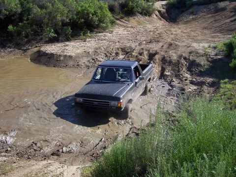 1991 Ford Bronco - A dark pickup truck navigates a muddy off-road trail, partially submerged in water.