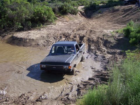 1991 Ford Bronco - A dark pickup truck navigates a challenging muddy terrain, with another vehicle visible in the background.