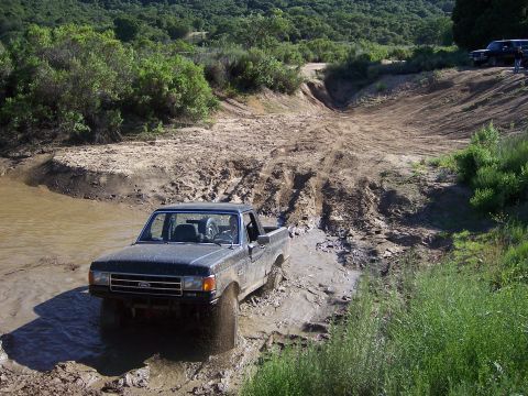 1991 Ford Bronco - A dark-colored Ford pickup truck navigates a muddy off-road trail with water and thick mud.
