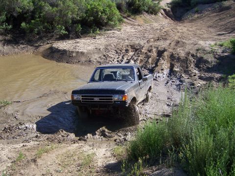 1991 Ford Bronco - A Ford truck navigates a muddy off-road trail with water obstacles.