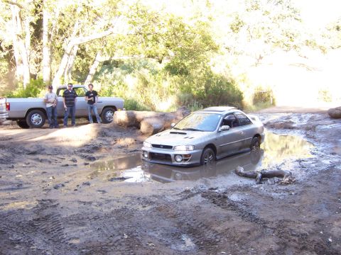 1991 Ford Bronco - A silver Subaru Impreza is stuck in a large mud puddle, while a pickup truck and three men stand by.