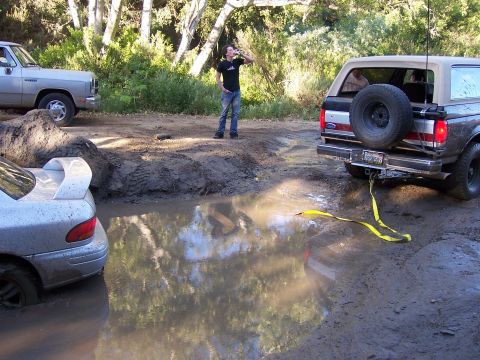 1991 Ford Bronco - Two vehicles are stuck in a muddy puddle, with one attempting to tow the other out.