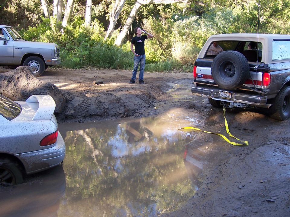 Ford Bronco pulling a Subaru out of a mud pit. A person stands nearby drinking