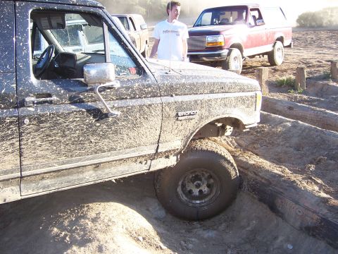 1991 Ford Bronco - Two Ford Broncos navigate an off-road course covered in mud and logs.