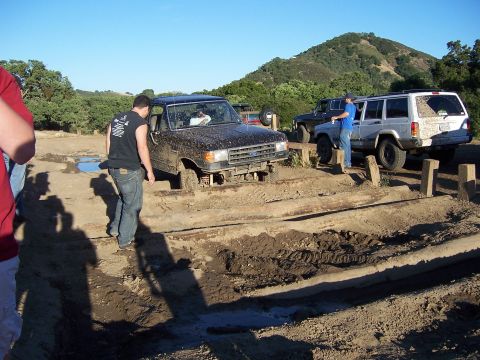 1991 Ford Bronco - Several off-road vehicles navigate a muddy course with wooden obstacles under a clear blue sky.