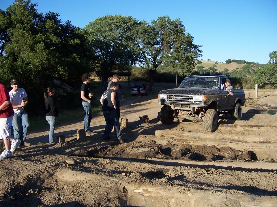 Modified Ford pickup truck with lifted suspension driving over dirt obstacles. Several people stand watching