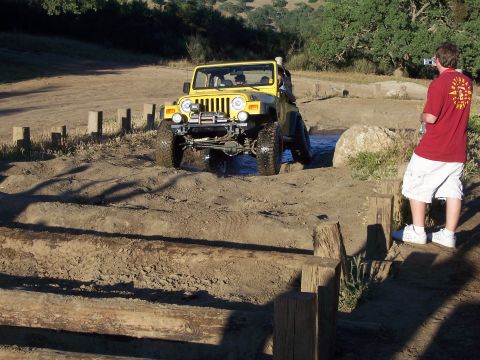 1991 Ford Bronco - A yellow Jeep Wrangler tackles a muddy off-road course, with a person filming in the foreground.