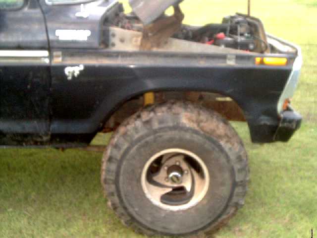 Front fender and wheel of a black Ford truck with hood open, showing engine parts. Yellow turn signal visible