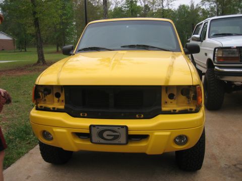 1996 Ford Bronco - A yellow Ford Ranger pickup truck is parked in front of another vehicle, showing its front end with missing headlights.