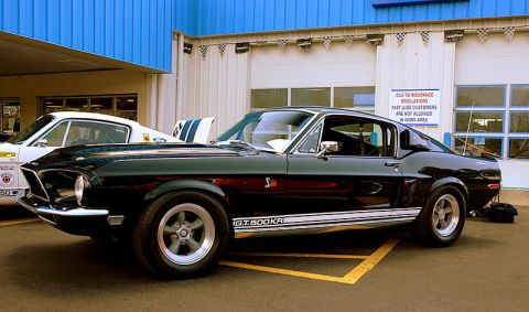 1968 Shelby GT500 - A black 1968 Shelby GT500KR Mustang is parked next to a white Mustang, with a sign on the building behind them.