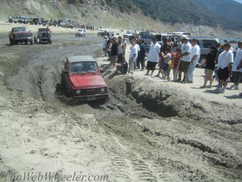1987 Suzuki Samurai - A red 1987 Suzuki Samurai tackles a challenging muddy terrain while onlookers observe the off-road action.