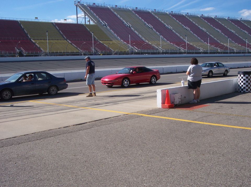 Red Ford Probe coupe on a racetrack with empty grandstands, near other cars and spectators