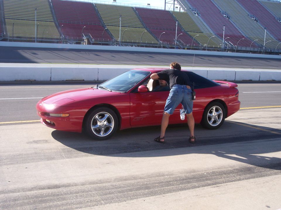 Red Ford Probe GT on a racetrack pit lane with a man leaning into the driver's window, grandstands behind