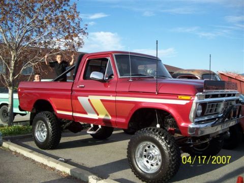 1976 Ford F-250 - A lifted red Ford F-250 pickup truck is shown with two people in the bed under a blue sky.