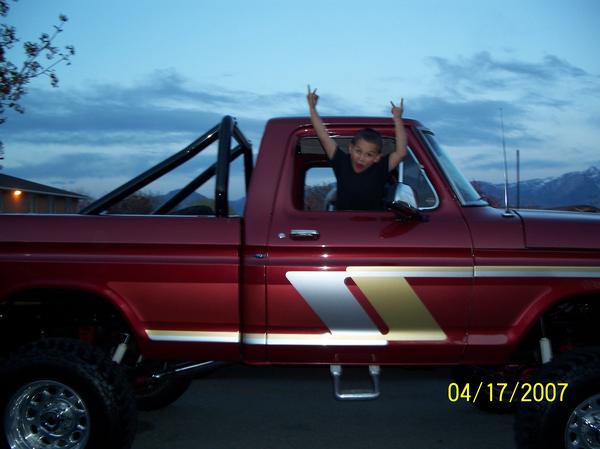 1976 Ford F-250 pickup truck, side view, with a boy in the driver's seat making rock-on hand gestures. The truck is red with silver and gold stripe decals on the door