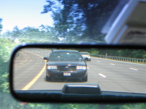 1991 Ford LTD Crown Victoria - A black sedan is seen in the rear-view mirror, driving on a highway with another vehicle ahead.