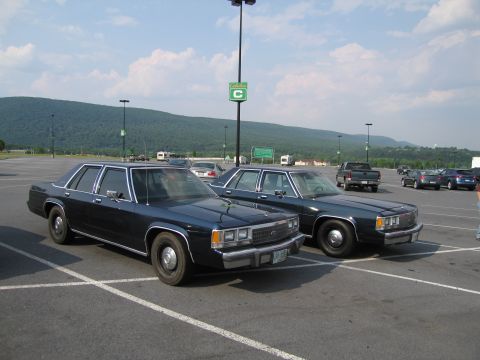1991 Ford LTD Crown Victoria - Two dark sedans, identified as 1991 Ford Crown Victoria LTDs, are parked side-by-side in a parking lot with a mountainous backdrop.