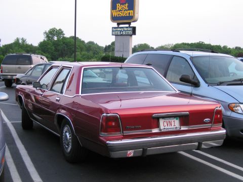 1991 Ford LTD Crown Victoria - A red 1991 Ford Crown Victoria LTD is parked next to a blue minivan, with a Best Western hotel sign in the background.