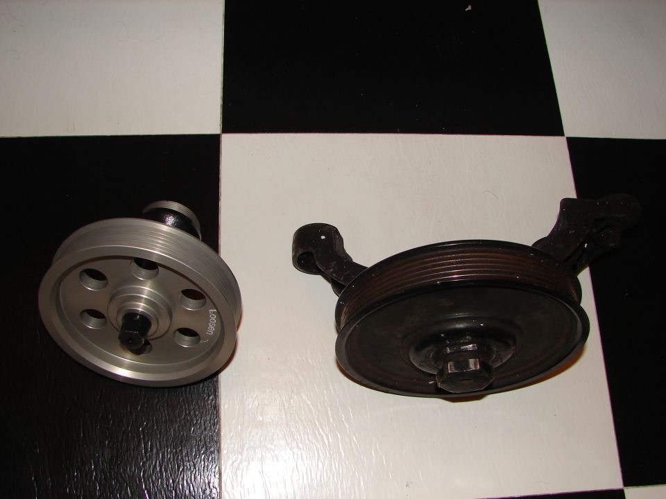 Two automotive pulleys on a black and white checkered surface; one is silver with holes, the other is black with a bracket
