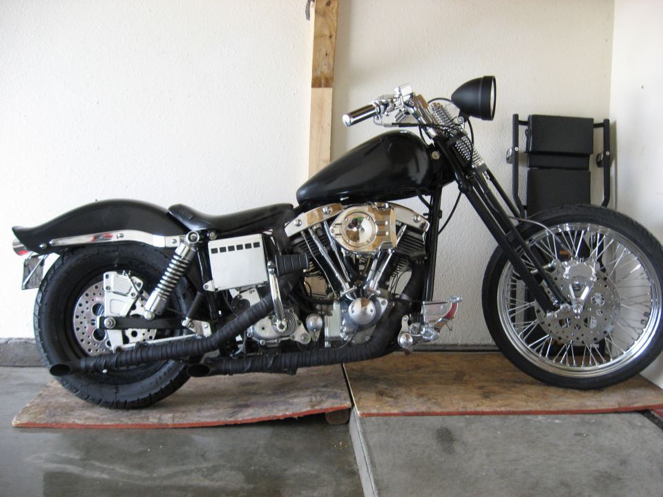 Black Harley Shovelhead chopper with wrapped exhaust and chrome spoked front wheel, parked in a garage, side view