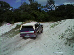 1986 Ford Bronco - A Ford Bronco navigates a sandy incline under a partly cloudy sky.