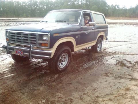 1986 Ford Bronco - A person drives a blue Ford Bronco through a muddy, watery area with trees in the background.