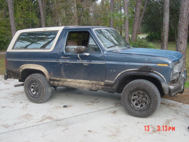 1986 Ford Bronco, blue, covered in mud, parked on concrete with trees in background